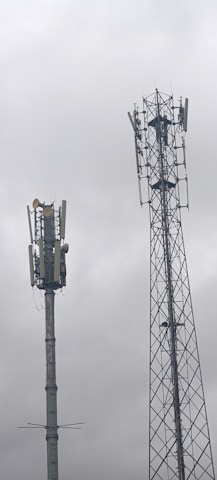 Two telecommunications towers are set against a cloudy sky. One tower has a solid cylindrical structure with various antennae attached, while the other tower has a lattice framework design with similar attachments.