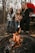 Three people sit around a campfire in a forested area. Two children are roasting marshmallows on sticks while an adult sits beside them on a picnic bench. A vibrant red tent is set up in the background among the leaf-covered ground and bare trees.