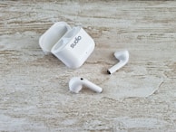 A pair of white wireless earbuds and an open charging case are placed on a textured wooden surface. The earbuds have a sleek design, and one of them is slightly off-angle compared to the other.