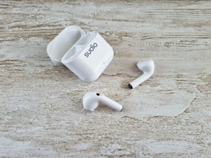 A pair of white wireless earbuds and an open charging case are placed on a textured wooden surface. The earbuds have a sleek design, and one of them is slightly off-angle compared to the other.