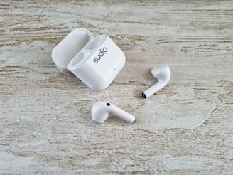 A pair of white wireless earbuds and an open charging case are placed on a textured wooden surface. The earbuds have a sleek design, and one of them is slightly off-angle compared to the other.