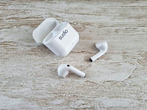 A pair of white wireless earbuds and an open charging case are placed on a textured wooden surface. The earbuds have a sleek design, and one of them is slightly off-angle compared to the other.