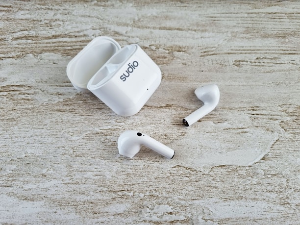 A pair of white wireless earbuds and an open charging case are placed on a textured wooden surface. The earbuds have a sleek design, and one of them is slightly off-angle compared to the other.