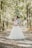 A couple embraces lovingly on a sun-dappled forest path. The bride wears a flowing white gown and has intricate detailing on her hair, while the groom is in a light-colored suit with a patterned bow tie. Tall trees on either side create a canopy of lush green leaves, casting soft shadows on the ground.