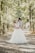 A couple embraces lovingly on a sun-dappled forest path. The bride wears a flowing white gown and has intricate detailing on her hair, while the groom is in a light-colored suit with a patterned bow tie. Tall trees on either side create a canopy of lush green leaves, casting soft shadows on the ground.