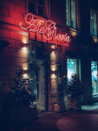 A warmly lit entrance to a building features a bright red neon sign with elegant cursive lettering. The door is flanked by two decorated Christmas trees adorned with lights and ornaments. The fa&ccedil;ade is constructed with large stones and illuminated by soft wall lights. The windows above and beside the entrance reflect a cozy interior ambiance.