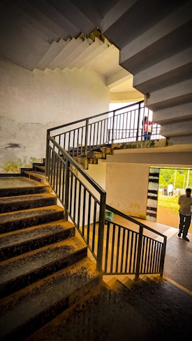 A multi-level staircase with metal railings, displaying a worn and slightly aged appearance. The view includes a section of stairs leading upward and downward, with a landing area. In the background, there are two people: one standing near a railing and one walking outside. The lighting creates contrasting shadows and highlights on the staircase surfaces.
