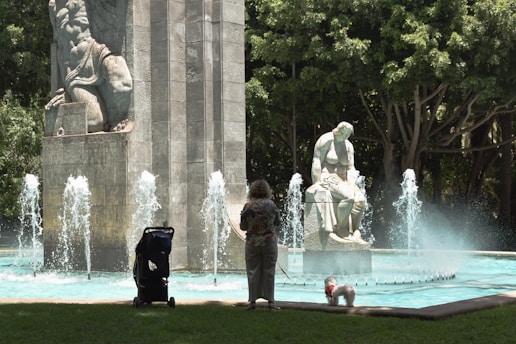 A person stands near a fountain with water jets shooting upwards, accompanied by a small white dog on a leash. The fountain features large stone statues, and a stroller is near the person. Surrounding the area are lush trees casting shade.