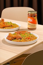 A table setting features plates of rice garnished with green peas and pieces of fried chicken. A jar of Gourmet Afrik Shito Pepper Sauce is prominently placed next to the plates. The scene is set on a light wooden table with neutral-colored chairs in the background.