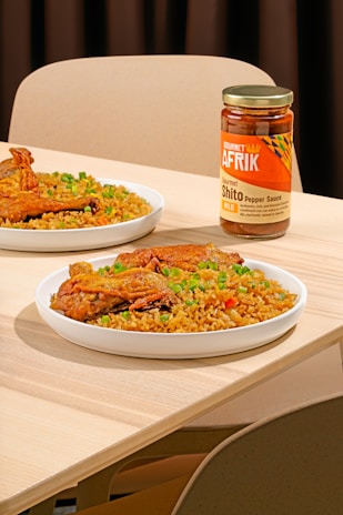 A table setting features plates of rice garnished with green peas and pieces of fried chicken. A jar of Gourmet Afrik Shito Pepper Sauce is prominently placed next to the plates. The scene is set on a light wooden table with neutral-colored chairs in the background.