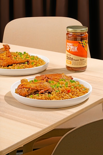 A table setting features plates of rice garnished with green peas and pieces of fried chicken. A jar of Gourmet Afrik Shito Pepper Sauce is prominently placed next to the plates. The scene is set on a light wooden table with neutral-colored chairs in the background.