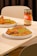 A table setting features plates of rice garnished with green peas and pieces of fried chicken. A jar of Gourmet Afrik Shito Pepper Sauce is prominently placed next to the plates. The scene is set on a light wooden table with neutral-colored chairs in the background.