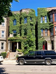 Two adjacent residential buildings with facades covered in lush green ivy. The structures are made of stone with large windows, and there is a black SUV parked on the street in front.