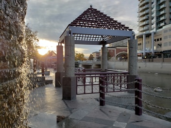 A riverside scene with a pavilion constructed from stone pillars and a wooden roof. Water cascades down a textured wall to the left as the sun sets on the horizon, casting a warm glow. A river runs beside modern buildings, creating a contrast between nature and urban development.