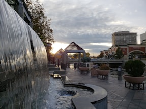 A cascading waterfall is seen in an urban setting, with water flowing down a vertical surface into a lower pool. The background features a modern cityscape with tall buildings, a pedestrian bridge, and geometric structures. Large planter pots with trees and shrubs line the paved walkway. The scene is under a cloudy sky with the sun partially visible on the horizon, casting a soft glow.