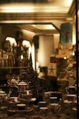 A cozy, dimly-lit shop display showcases a collection of intricately designed teapots, teacups, and saucers. The arrangement includes various patterns and colors, all set against a backdrop of shelves filled with an assortment of items. A decorative, slender tree with sparkling lights adds a festive touch to the scene.