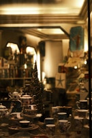 A cozy, dimly-lit shop display showcases a collection of intricately designed teapots, teacups, and saucers. The arrangement includes various patterns and colors, all set against a backdrop of shelves filled with an assortment of items. A decorative, slender tree with sparkling lights adds a festive touch to the scene.