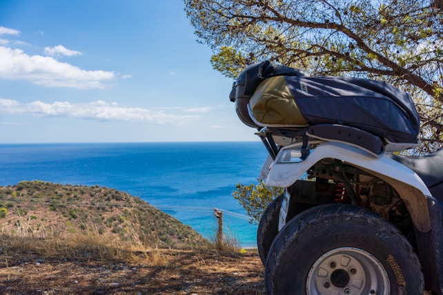A scenic seaside landscape features a four-wheeler parked on a dirt path. The view includes a vast ocean under a bright blue sky, with a few scattered clouds. In the foreground, dry grass and shrubbery contrast with the vibrant blue of the water. A sturdy tree with sparse foliage frames the right side of the scene.