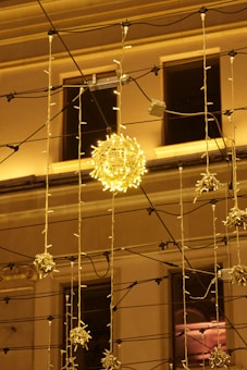 A warmly lit building facade with intricate decorative light fixtures, including a glowing orb surrounded by string lights and electric wires.