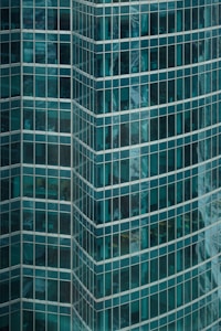 A modern glass skyscraper with reflective windows, showing partial views of interior office spaces and external reflections. The building features a geometric pattern formed by rows of rectangular windows.