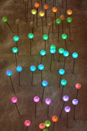 Multiple colorful glowing spheres are mounted on thin black sticks, scattered across a dark surface. The spheres are illuminated in various shades, including green, blue, pink, and orange, creating a vibrant and visually stimulating pattern.