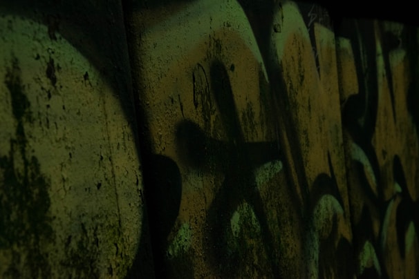Dark graffiti art covers a concrete wall, with abstract shapes and shadowy textures. The muted greenish tones blend with darker shadows, creating a mysterious and gritty atmosphere.