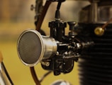 A close-up view of a mechanical component, likely part of an engine or a machine. A prominent, shiny metallic cylinder with a mesh front is connected to various black and silver parts and tubes. The background is out of focus, suggesting this is the focus of the image.