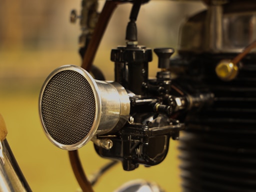 A close-up view of a mechanical component, likely part of an engine or a machine. A prominent, shiny metallic cylinder with a mesh front is connected to various black and silver parts and tubes. The background is out of focus, suggesting this is the focus of the image.