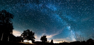 An expansive night sky filled with countless stars and a visible Milky Way galaxy. In the foreground, a brightly lit tent is set up amidst silhouettes of trees and grassy terrain.