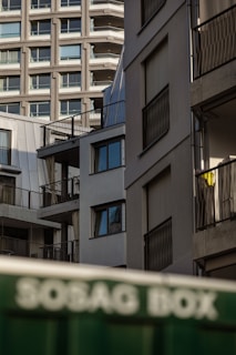A view of modern residential buildings characterized by balconies and large windows. The architecture includes straight lines and a minimalist design, while a green container with partly visible text appears at the bottom.