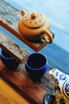 A yellow teapot with Chinese characters rests on a rough wooden shelf. Below the shelf, there are two dark blue cups and part of another teapot with a floral design. The background is slightly blurred and appears to be a window.