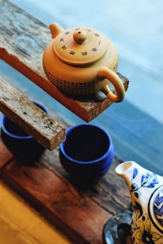 A yellow teapot with Chinese characters rests on a rough wooden shelf. Below the shelf, there are two dark blue cups and part of another teapot with a floral design. The background is slightly blurred and appears to be a window.