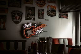 A vintage-themed room with various retro-style posters and memorabilia on the walls. A neon sign depicting a guitar with the words 'Rock & Roll' is prominently displayed. The seating consists of red and white cushioned booths, and there are condiments on the tables.