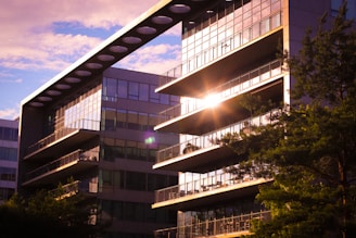 A modern building with glass windows reflecting sunlight, surrounded by trees. The architecture features a striking horizontal beam with circular cutouts along the top, adding a contemporary design element.