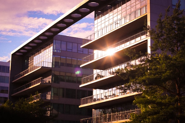 A modern building with glass windows reflecting sunlight, surrounded by trees. The architecture features a striking horizontal beam with circular cutouts along the top, adding a contemporary design element.