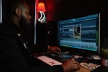 A person working at a music production setup with a large monitor displaying music editing software. The room has dim lighting with a red lamp casting a warm glow. Various studio equipment and accessories, such as a keyboard and mixing console, are present.