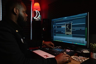 A person working at a music production setup with a large monitor displaying music editing software. The room has dim lighting with a red lamp casting a warm glow. Various studio equipment and accessories, such as a keyboard and mixing console, are present.
