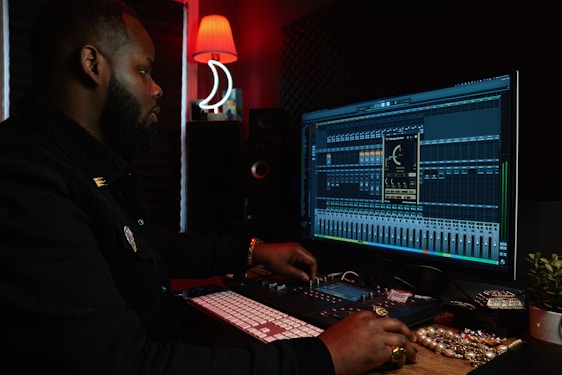 A person working at a music production setup with a large monitor displaying music editing software. The room has dim lighting with a red lamp casting a warm glow. Various studio equipment and accessories, such as a keyboard and mixing console, are present.