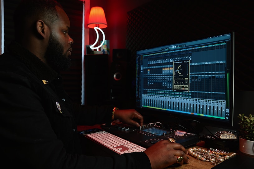 A person working at a music production setup with a large monitor displaying music editing software. The room has dim lighting with a red lamp casting a warm glow. Various studio equipment and accessories, such as a keyboard and mixing console, are present.