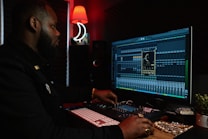 A person working at a music production setup with a large monitor displaying music editing software. The room has dim lighting with a red lamp casting a warm glow. Various studio equipment and accessories, such as a keyboard and mixing console, are present.