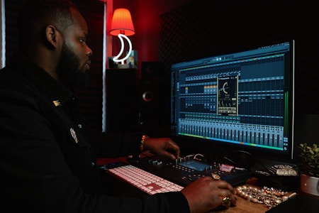A person working at a music production setup with a large monitor displaying music editing software. The room has dim lighting with a red lamp casting a warm glow. Various studio equipment and accessories, such as a keyboard and mixing console, are present.