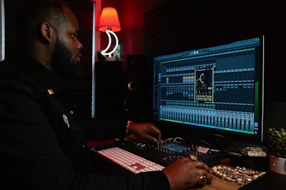A person working at a music production setup with a large monitor displaying music editing software. The room has dim lighting with a red lamp casting a warm glow. Various studio equipment and accessories, such as a keyboard and mixing console, are present.