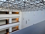 A modern interior space with clean architectural lines and multiple levels visible. The ceiling features an intricate lattice framework, allowing light to filter through. Walls are pristine white with small rectangular windows, and the floors have warm lighting near the doors.