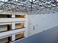 A modern interior space with clean architectural lines and multiple levels visible. The ceiling features an intricate lattice framework, allowing light to filter through. Walls are pristine white with small rectangular windows, and the floors have warm lighting near the doors.
