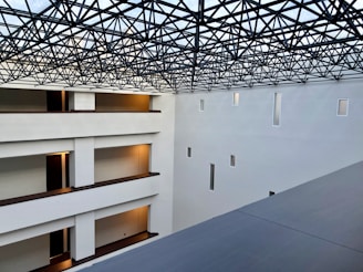 A modern interior space with clean architectural lines and multiple levels visible. The ceiling features an intricate lattice framework, allowing light to filter through. Walls are pristine white with small rectangular windows, and the floors have warm lighting near the doors.