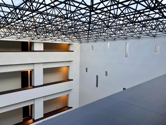 A modern interior space with clean architectural lines and multiple levels visible. The ceiling features an intricate lattice framework, allowing light to filter through. Walls are pristine white with small rectangular windows, and the floors have warm lighting near the doors.