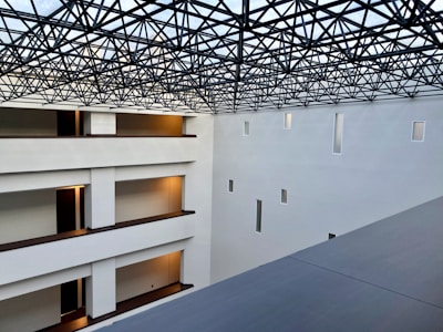 A modern interior space with clean architectural lines and multiple levels visible. The ceiling features an intricate lattice framework, allowing light to filter through. Walls are pristine white with small rectangular windows, and the floors have warm lighting near the doors.