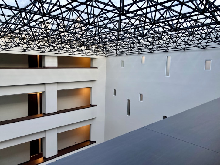 A modern interior space with clean architectural lines and multiple levels visible. The ceiling features an intricate lattice framework, allowing light to filter through. Walls are pristine white with small rectangular windows, and the floors have warm lighting near the doors.