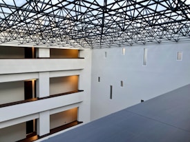 A modern interior space with clean architectural lines and multiple levels visible. The ceiling features an intricate lattice framework, allowing light to filter through. Walls are pristine white with small rectangular windows, and the floors have warm lighting near the doors.