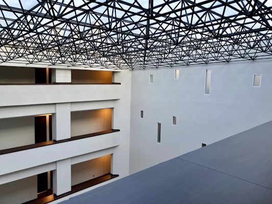 A modern interior space with clean architectural lines and multiple levels visible. The ceiling features an intricate lattice framework, allowing light to filter through. Walls are pristine white with small rectangular windows, and the floors have warm lighting near the doors.